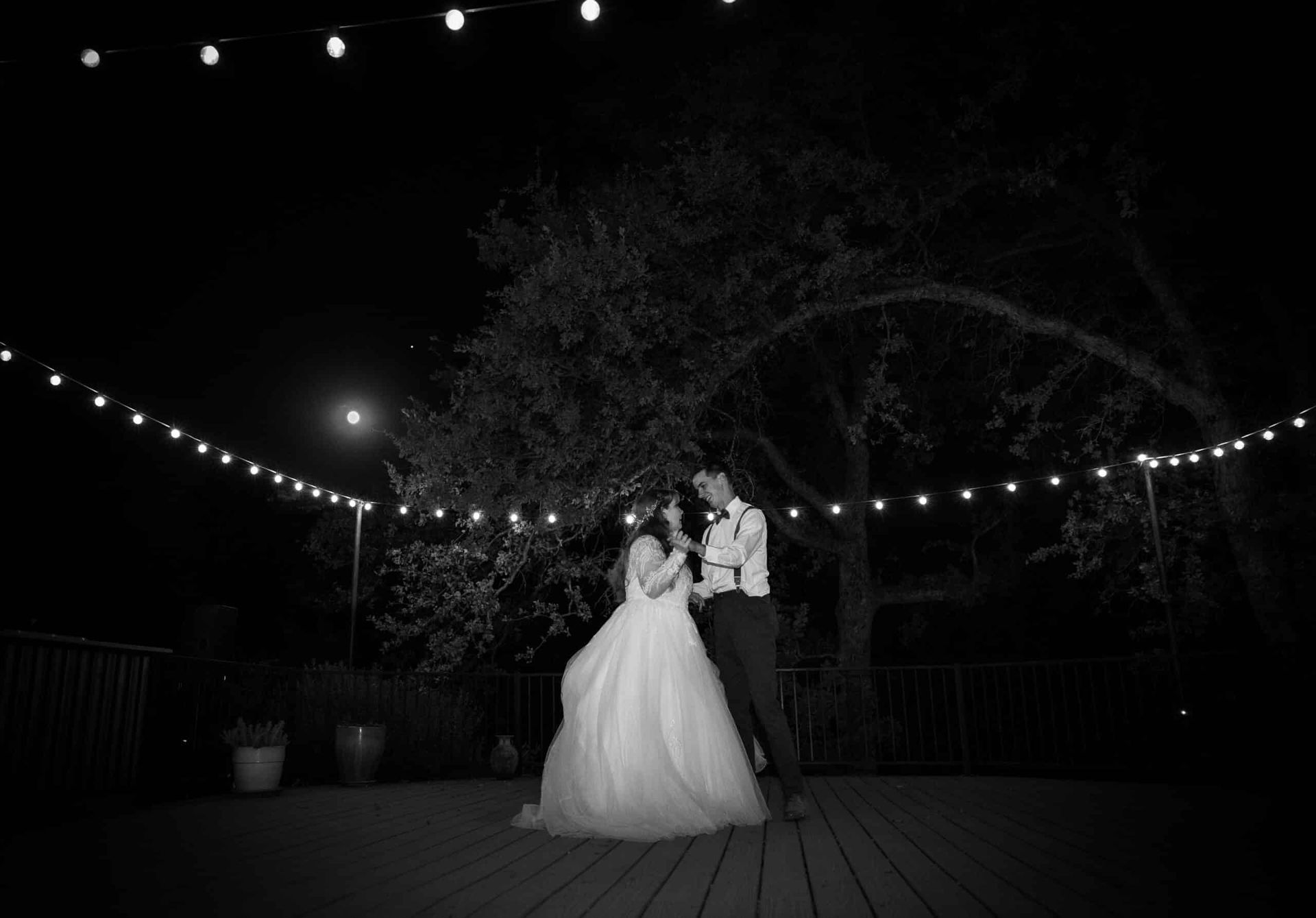 A bride and groom are standing on a deck at night under a string of lights.