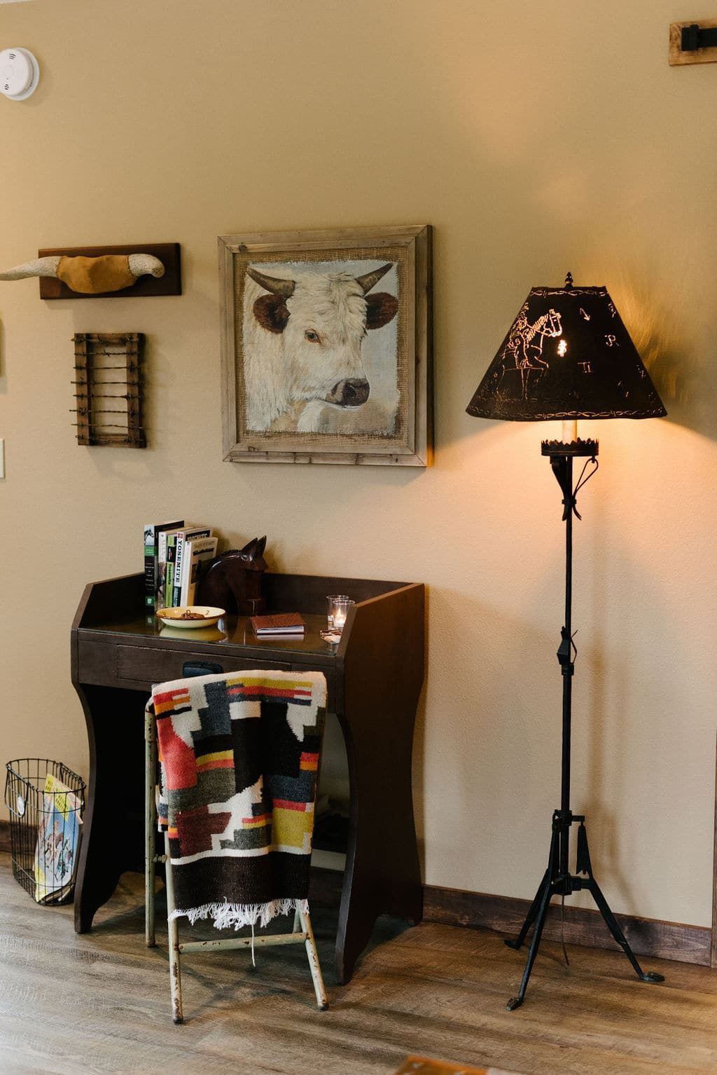 A room with a desk , chair , lamp and a painting of a cow on the wall.