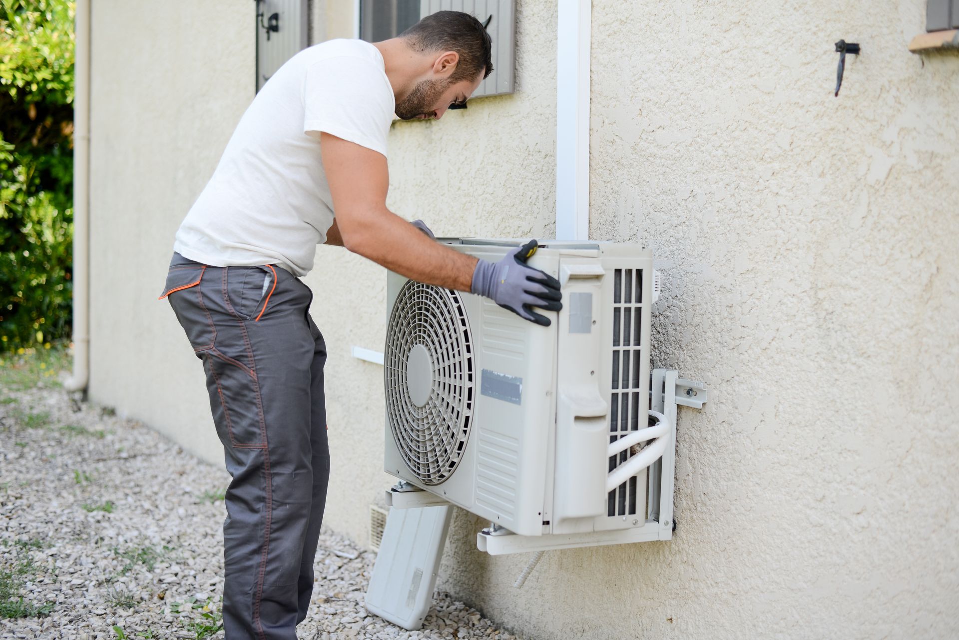 Man repairing an outdoor air conditioner unit mounted on a wall, wearing gloves and leaning in to work.
