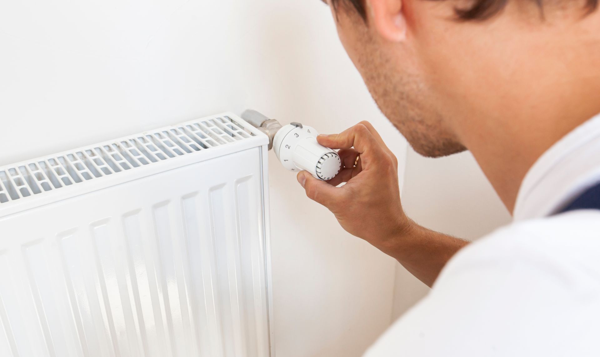 Man adjusting a white radiator thermostat knob in a bright room