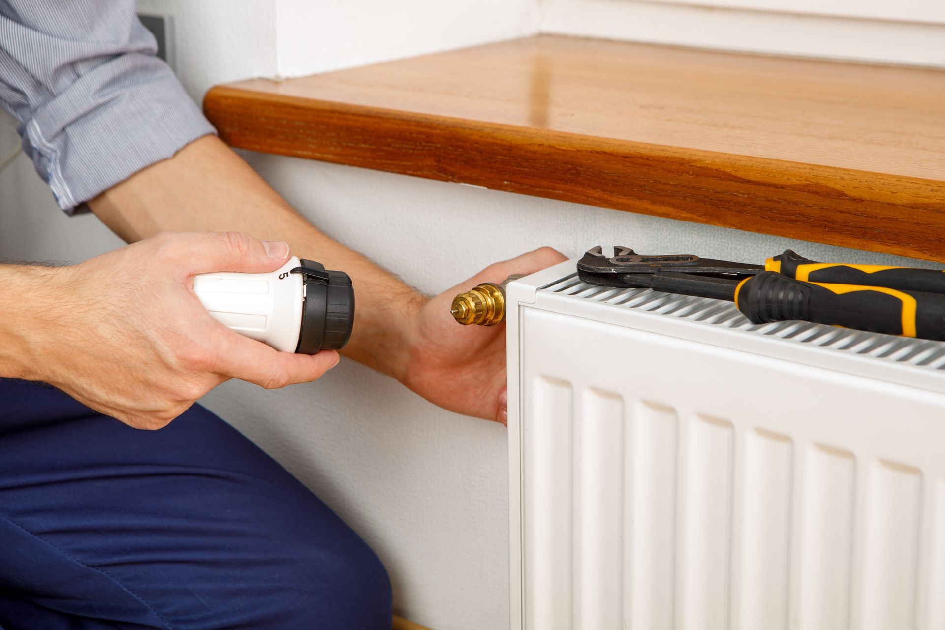 Hand holding a thermostatic radiator valve under a wooden shelf with tools nearby