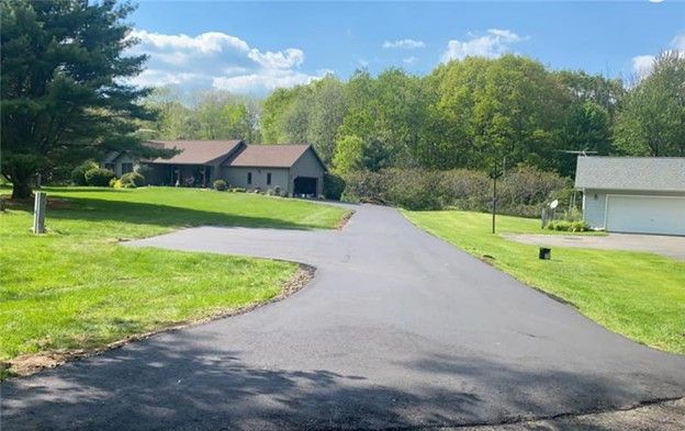Asphalt driveway curves toward a house with a garage, surrounded by green grass and trees under a blue sky.