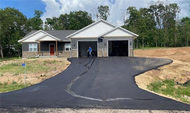 New asphalt driveway leading to a gray house with a red door and two-car garage; a person is working on the driveway.