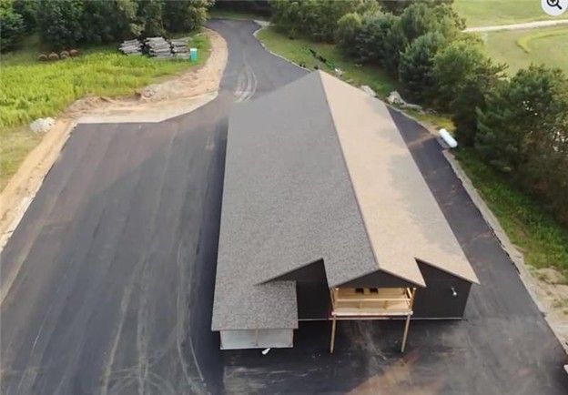 Aerial view of a long, dark-sided building with a light-colored roof and asphalt driveway in a grassy setting.