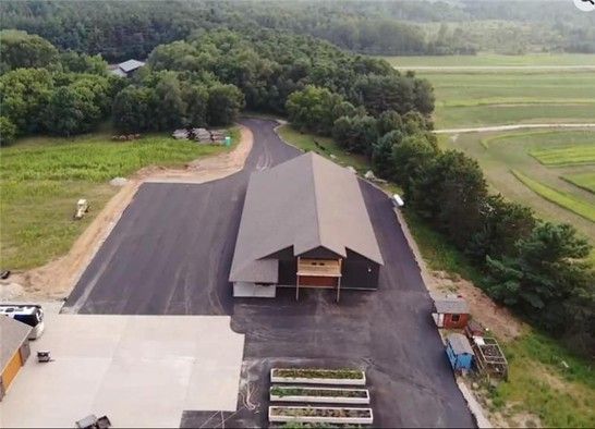 Aerial view of a large, dark-roofed building with a paved driveway and surrounding fields and trees.