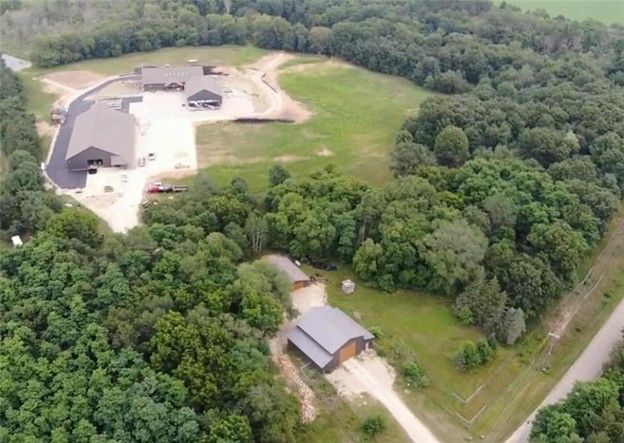 Aerial view of a rural property with buildings, a gravel driveway, and surrounding green trees.