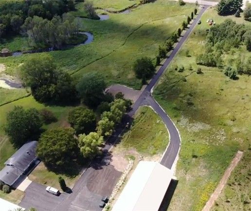 Aerial view: a paved road curves through green fields, trees, and a building; sunny day.