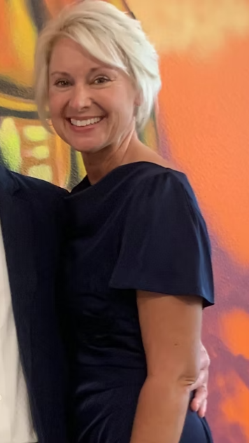 Woman with glasses smiles; teal and pink patterned blouse; blurred background.