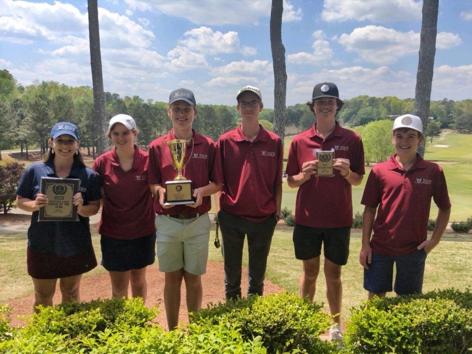 Five people in maroon shirts hold trophies outdoors on a sunny sports field.