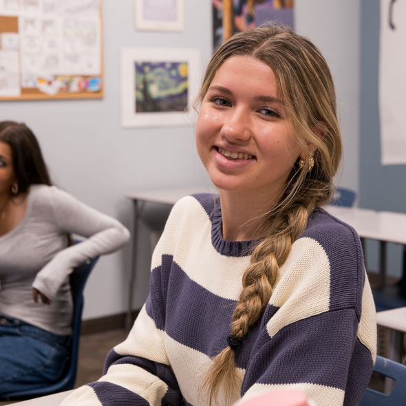 Smiling woman with a braid in a striped sweater seated in a classroom or office lounge.