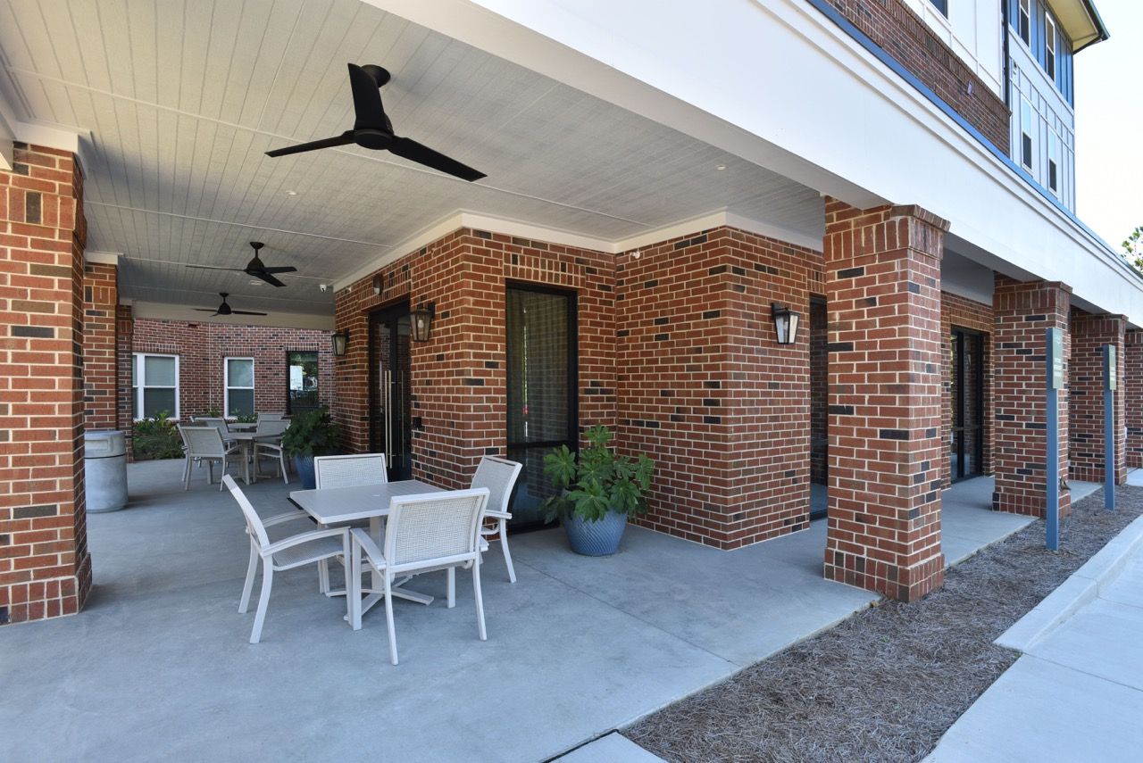 Covered brick patio with outdoor tables and chairs beneath a walkway along the building.