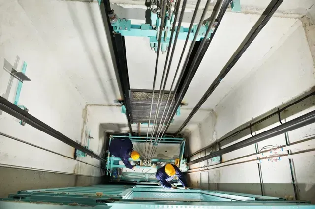 Dos trabajadores con cascos inspeccionan un hueco de ascensor profundo con cables y raíles.