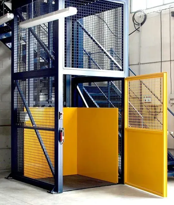 Montacargas industrial con marco azul y puerta de seguridad amarilla en el interior de una fábrica.