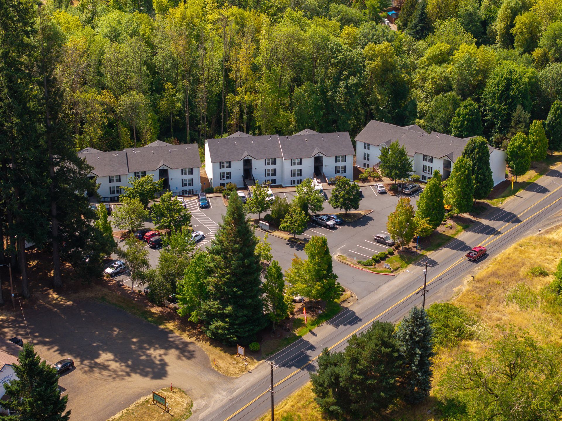 Aerial view of an apartment complex surrounded by trees and a parking lot next to a road with cars.