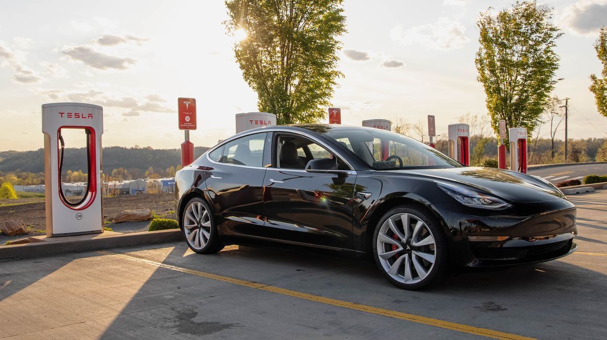 Black electric sedan parked at a Tesla charging station at sunset, with trees and city skyline in the background