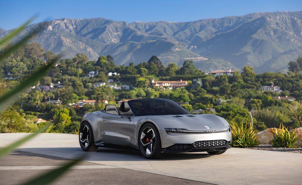 Silver convertible sports car parked on a road with mountains and greenery in the background