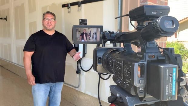Man standing indoors beside a professional video camera filming him in a hallway