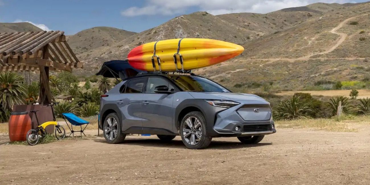 Gray SUV with a surfboard on the roof parked on a dirt road beside a thatched hut, hills in the background