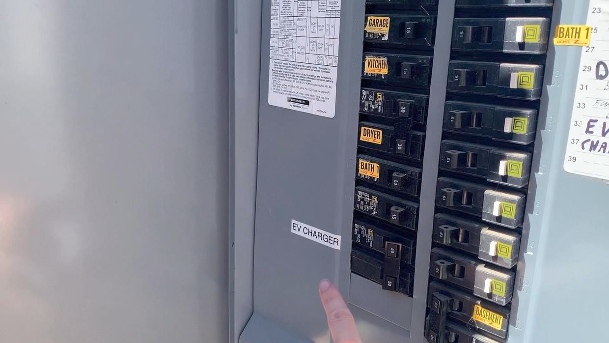 Person pointing at an electrical breaker panel with numbered switches and labels.