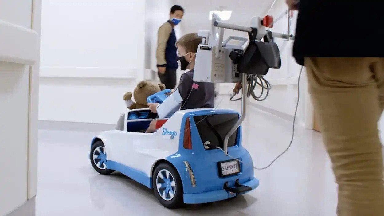 Child riding a blue toy car indoors while an adult stands nearby with a hand truck.