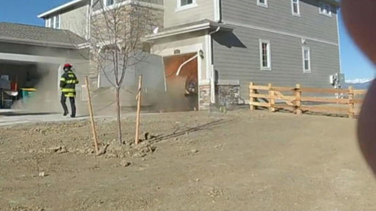 Worker in high-visibility gear stands outside a gray house near a garage and wooden fence on a dirt lot.