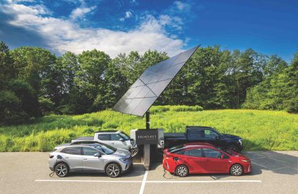 Two small cars parked by a solar panel on a grassy roadside under a blue sky