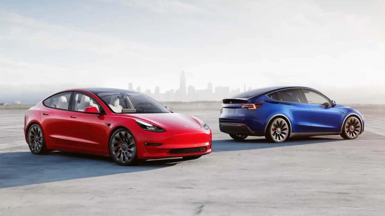 Two Tesla sedans, red and blue, parked on a rooftop with a city skyline in the background