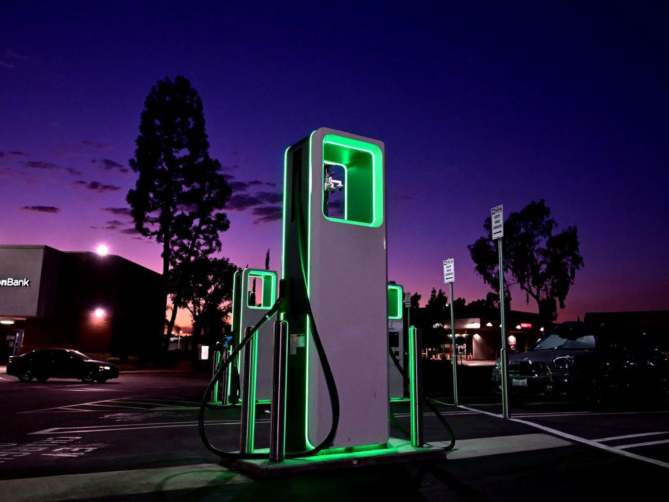 Green-lit EV charging stations at dusk in a parking lot under a purple sky.