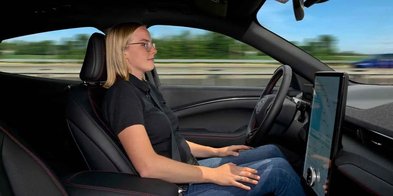 Woman driving a car, viewed from the passenger side, with a dashboard screen visible.