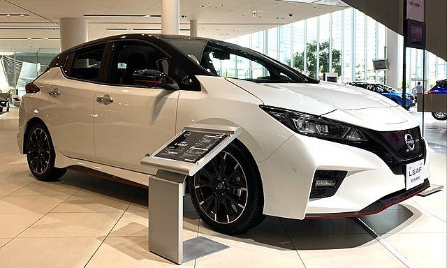 White hatchback car displayed in a showroom, viewed from the front-left, with a small info stand nearby