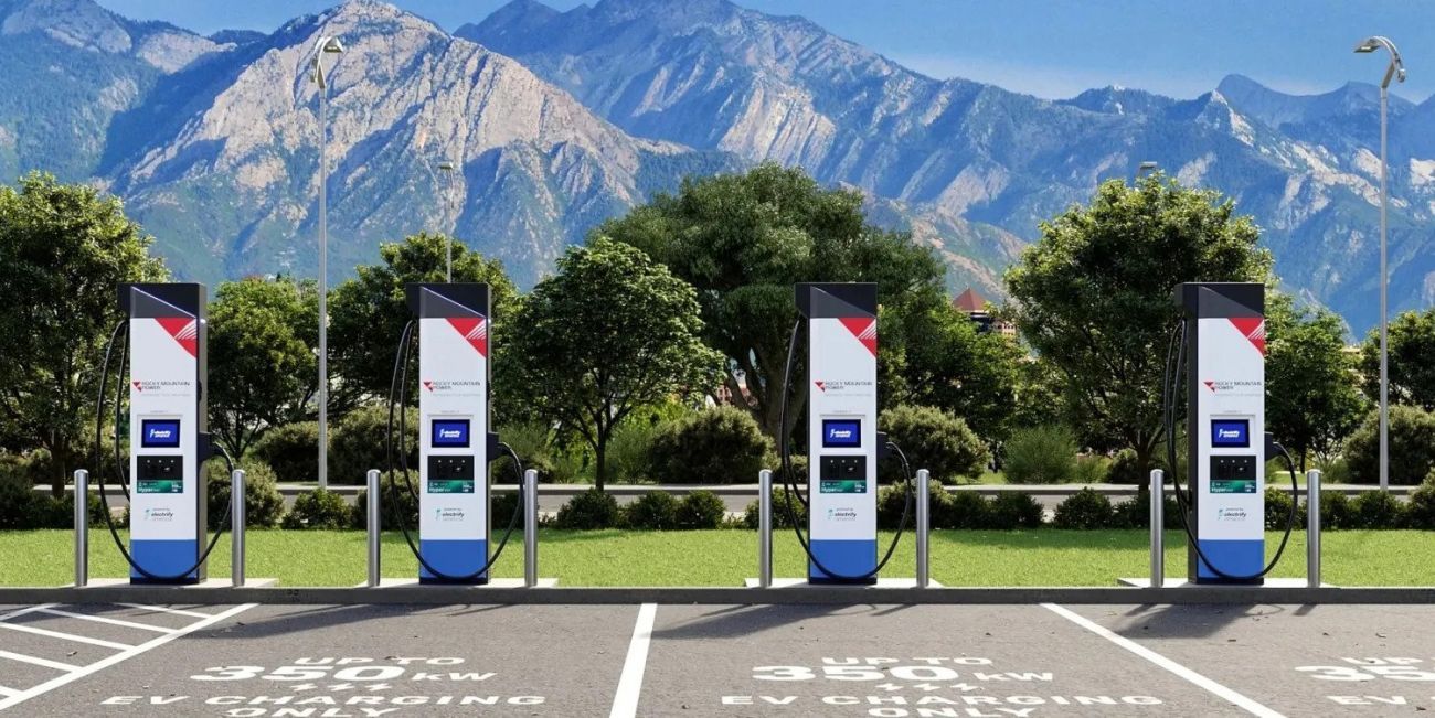 Four blue-and-white EV charging stations in a parking lot with mountains in the background