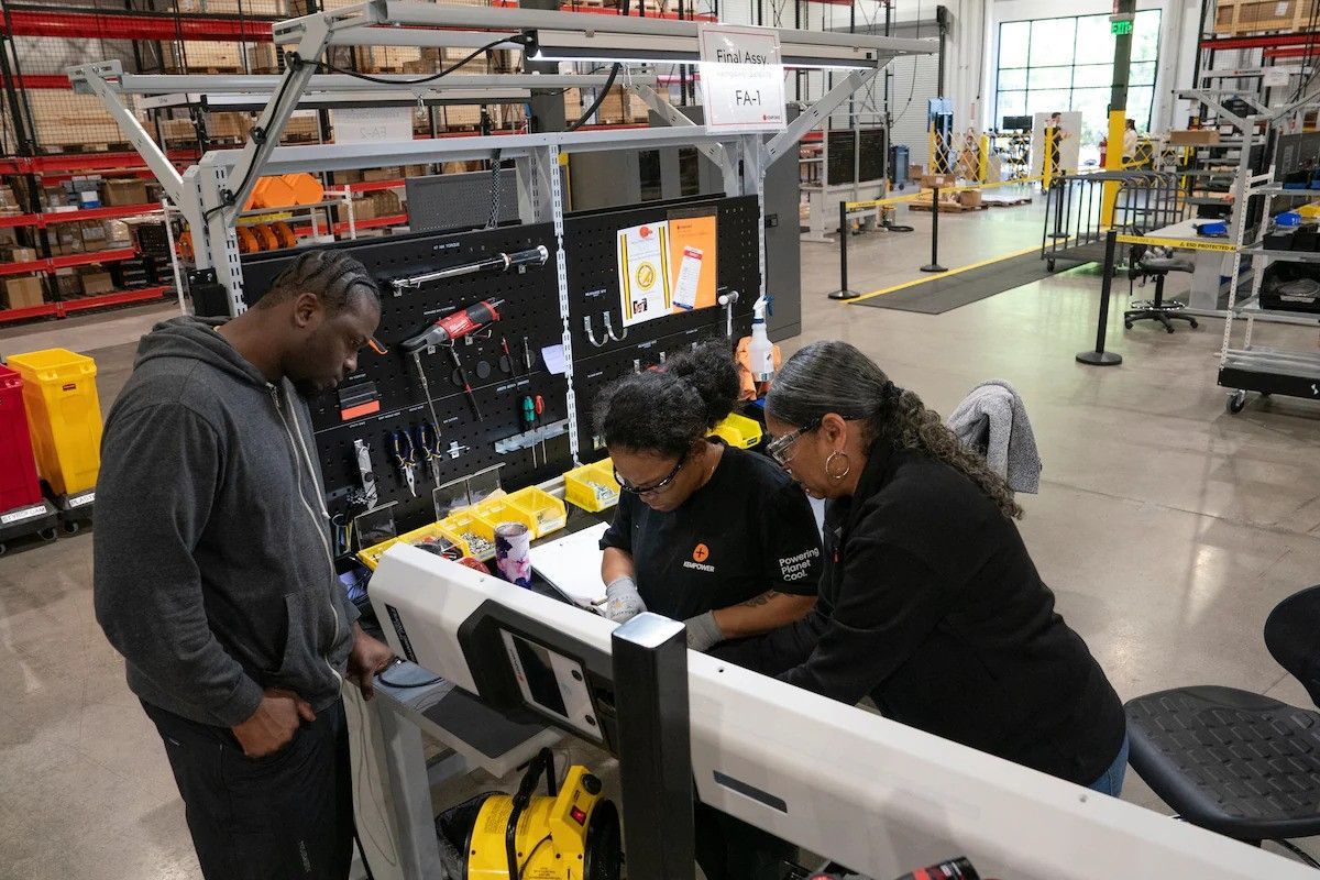 People collaborating at a workstation in a warehouse, with tools, monitors, and shelving in the background.