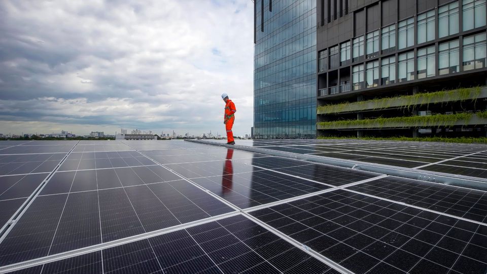 Une personne vêtue d’une tenue de travail orange sur un toit couvert de panneaux solaires, à côté d’un immeuble moderne en verre.