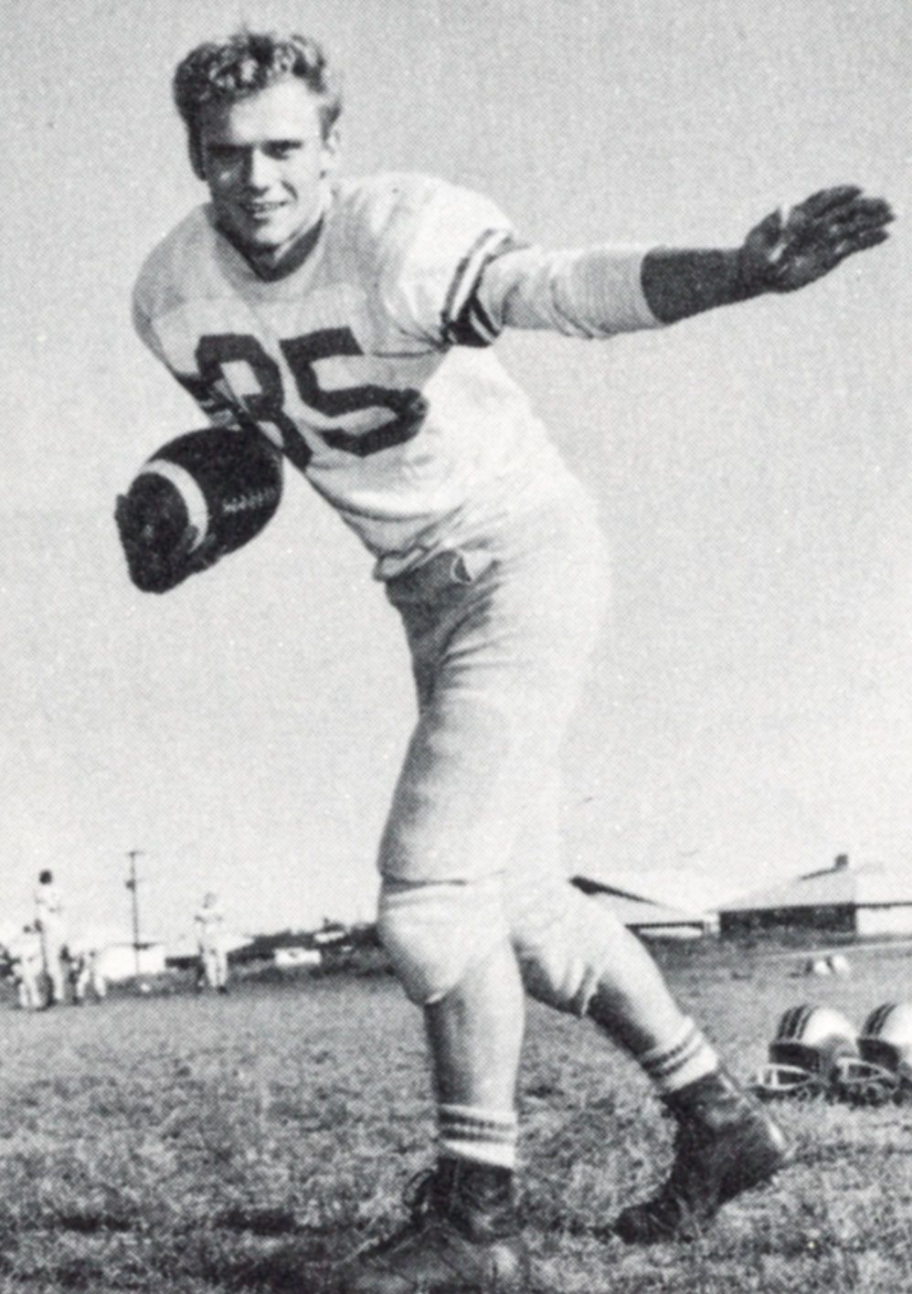 Smiling football player (#85), holding a football in one hand and with the other arm out, as if to block.