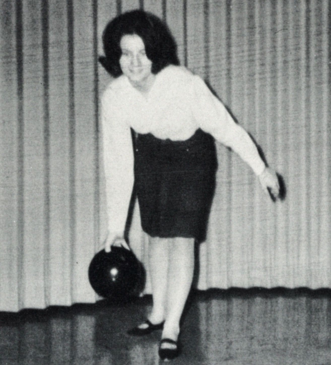 Female student in a skirt, posing with a bowling ball, as if she is ready to throw it down the lane.