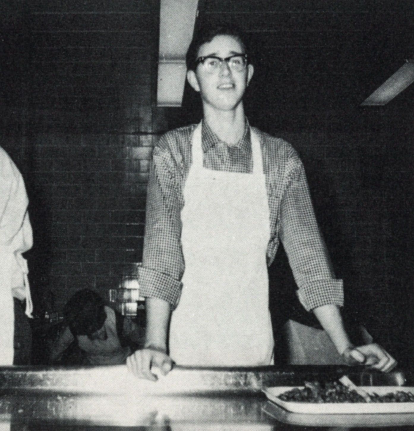 Smiling male student wearing an apron and standing behind a counter in the kitchen area