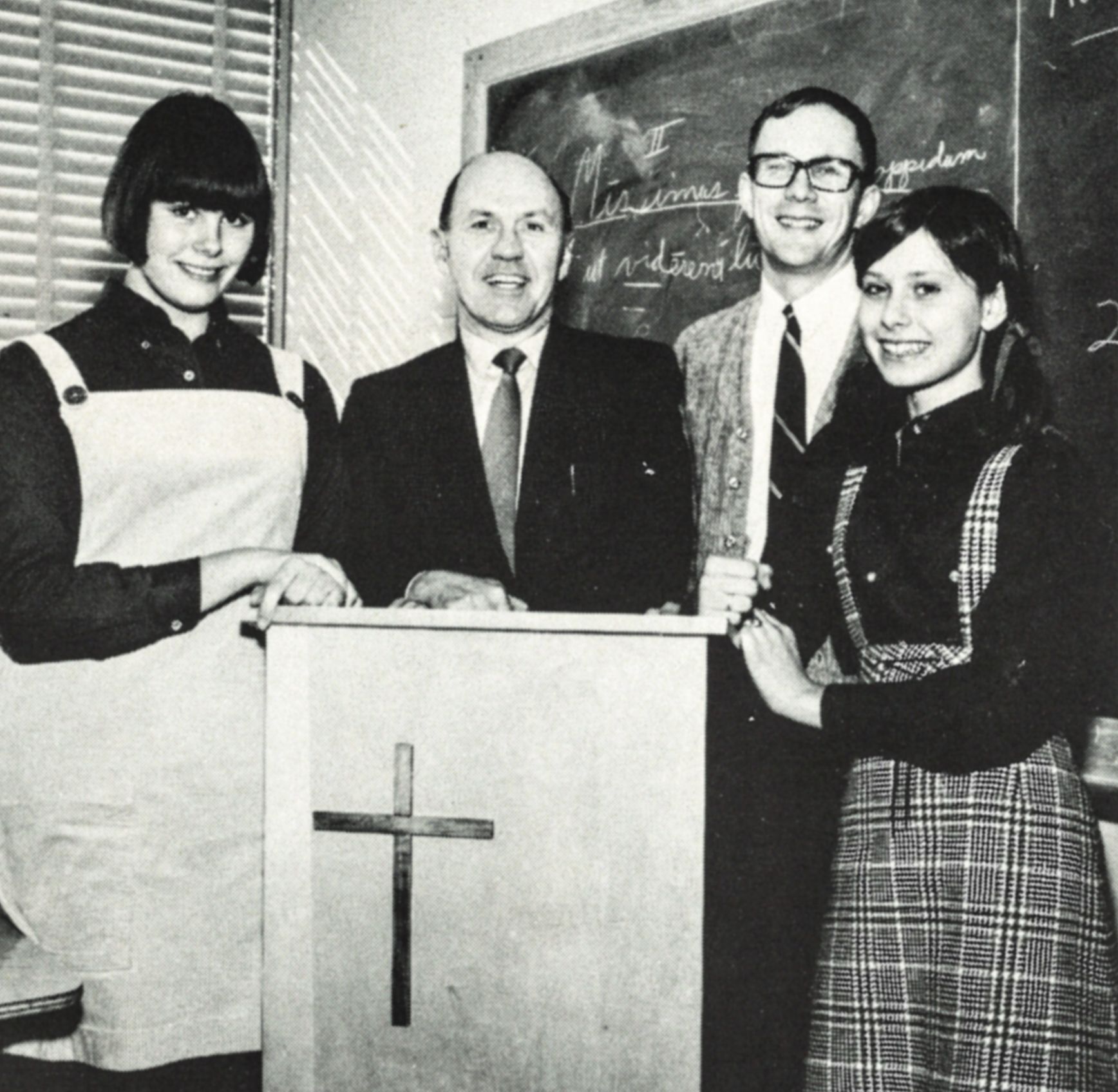 Students and teachers around a podium in a classroom. Chalkboard in the background