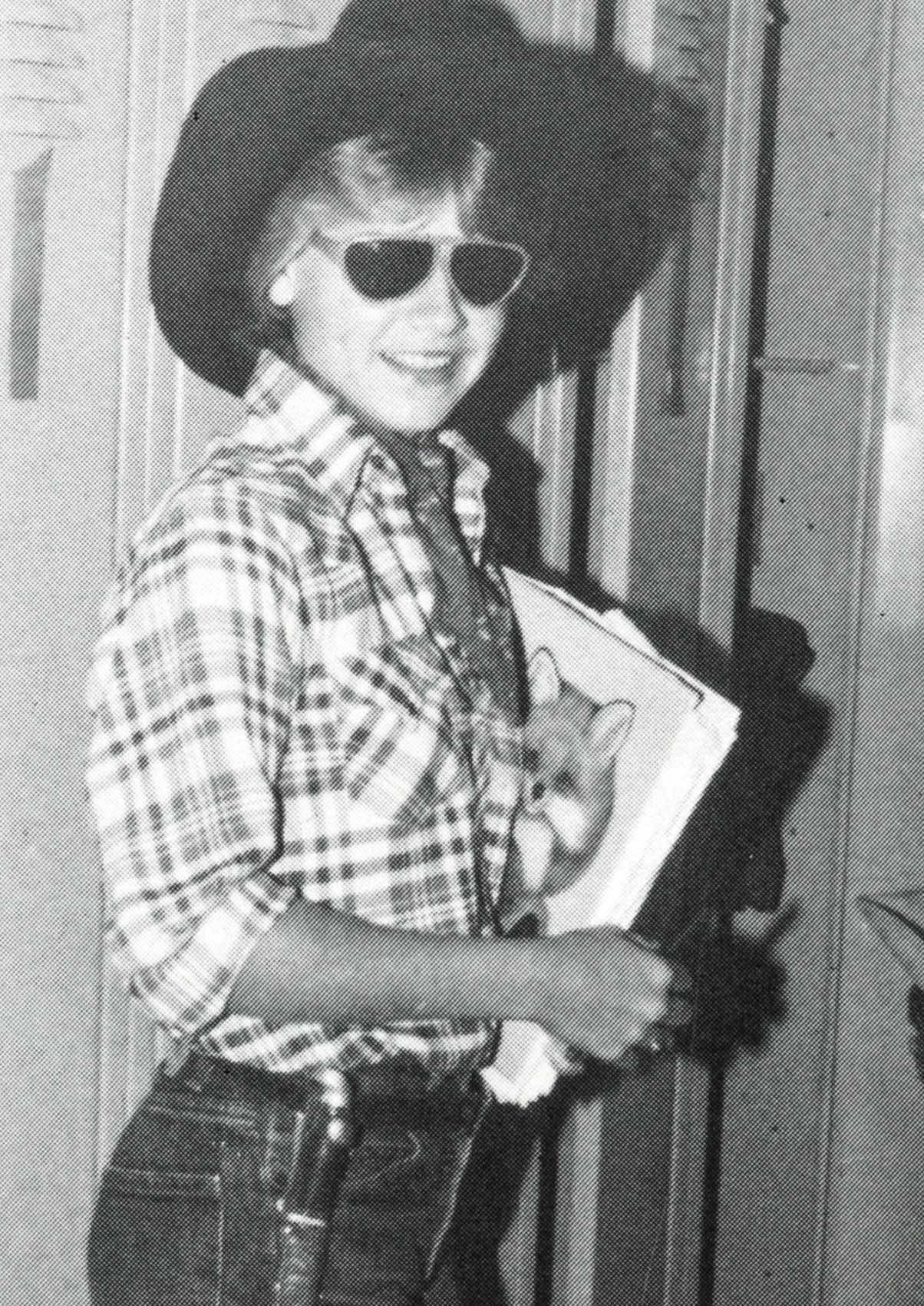 Female student in cowboy hat, cowboy shirt, and sunglasses, standing with books and smiling at the camera