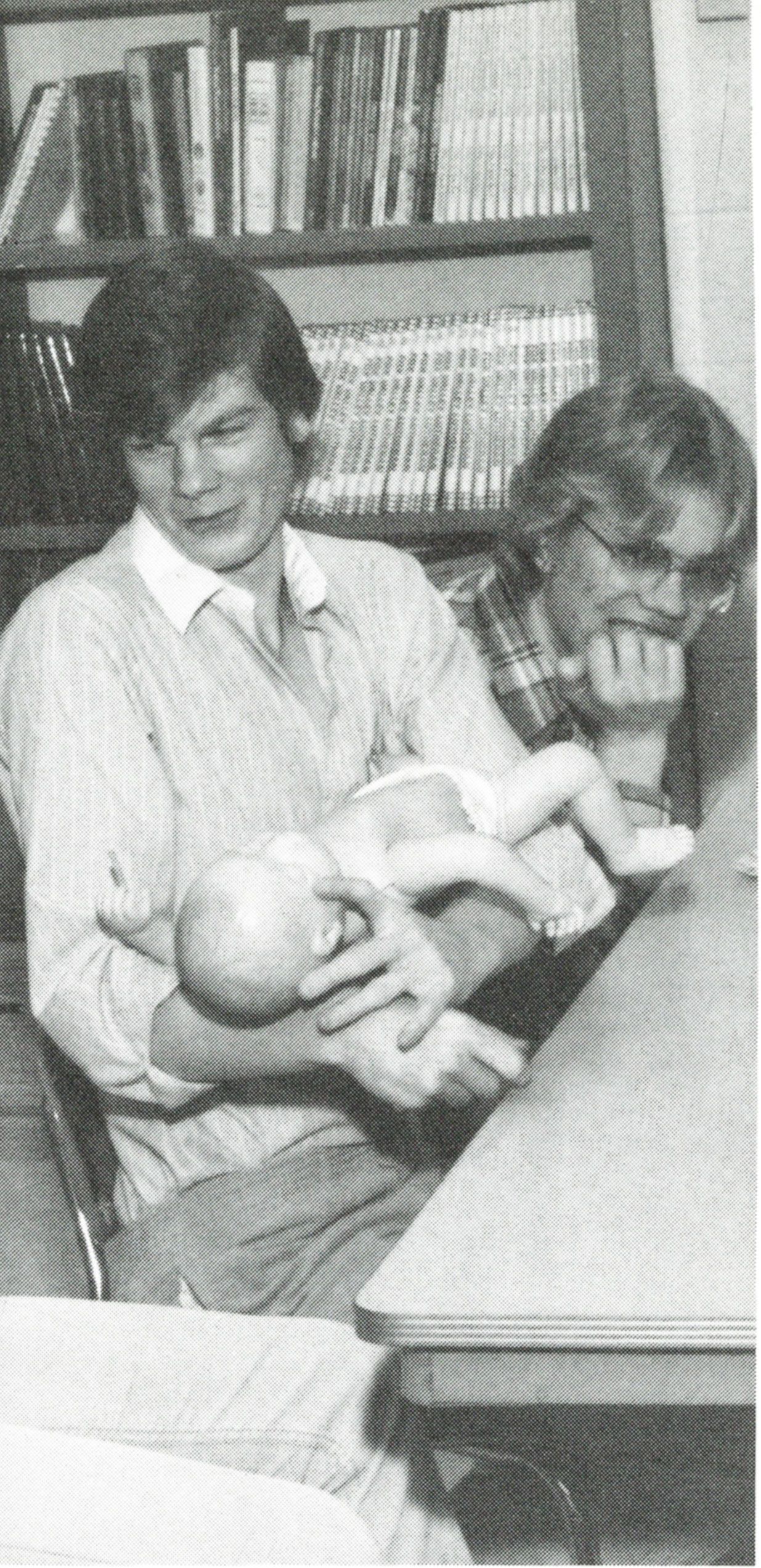 Two male students sitting at a table smiling, one is cradling a baby (it's a doll)