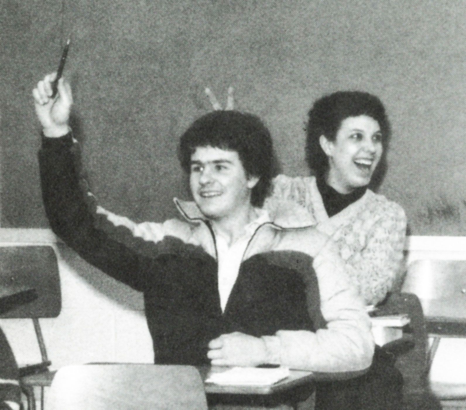 Two students sitting at desks. Male student is raising his hand. Female student is giving him rabbit ears and is laughing.