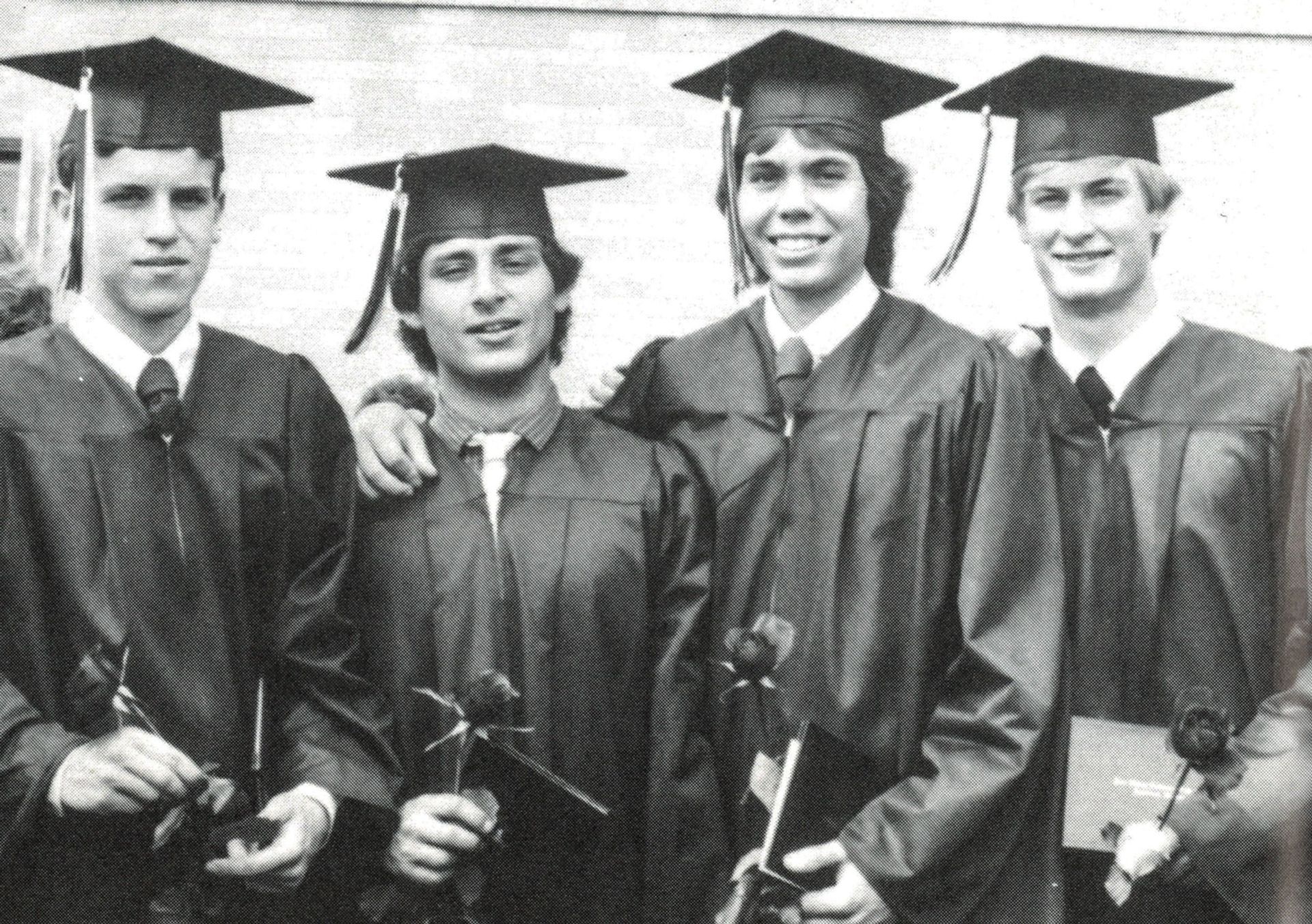 Four male students in cap and gown and each are holding a flower and their diplomas