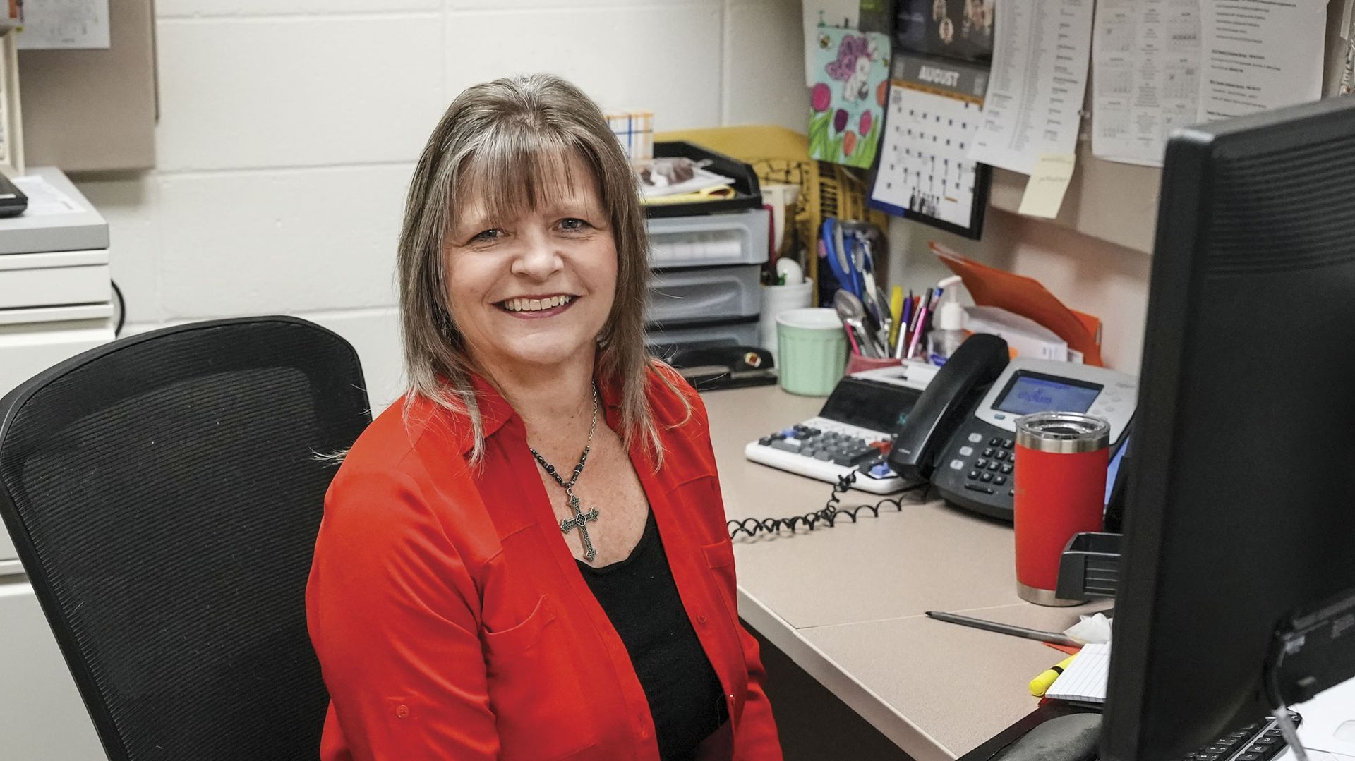 Krista in a red jacket, sitting at her desk and smiling