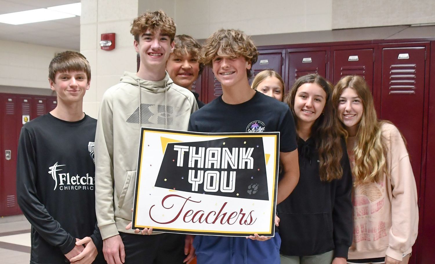 Group of seven students in the locker hallway, holding a sign that says