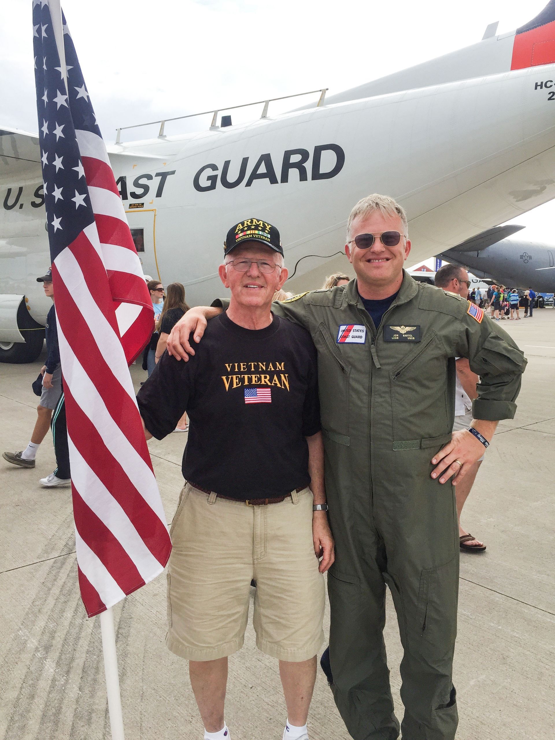 Jon and his father (who is holding a US flag), smiling and standing in front of a Coast Guard airplane