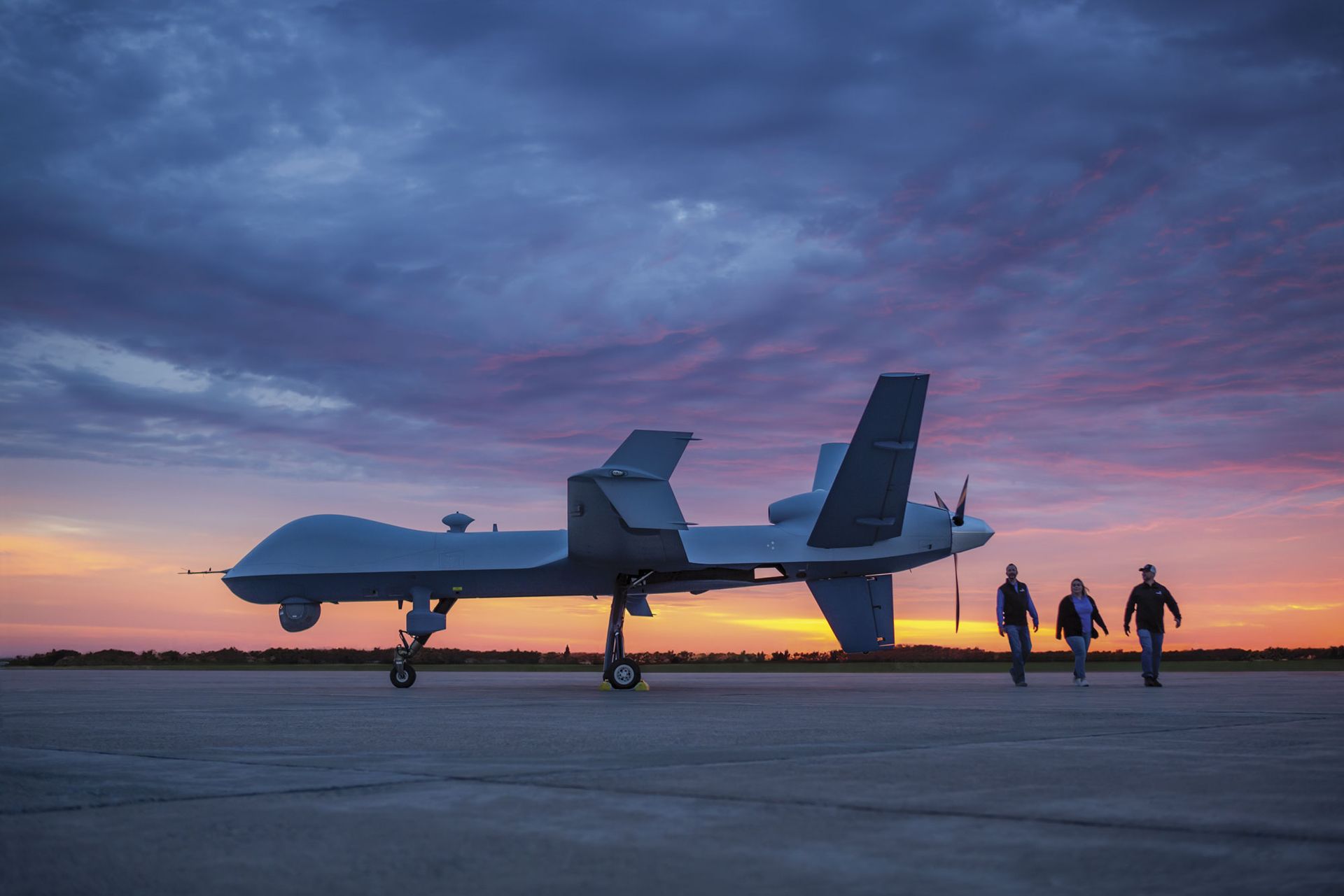 Large drone on tarmac with sunrise or sunset behind it and three people walking