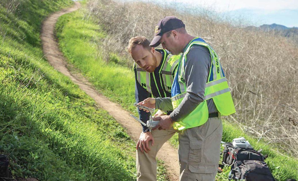 Aaron with another investigator on a grassy hillside, holding and discussing a piece of debris