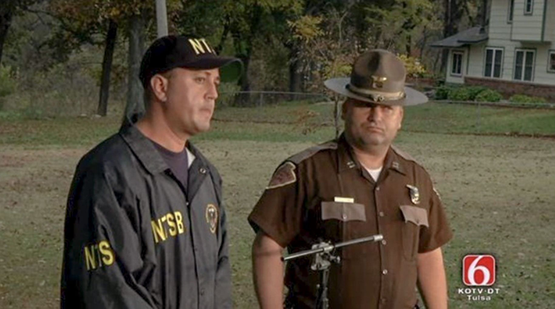 Aaron Sauer and local law enforcement officer on a TV screen in Tulsa. Woods and part of a house in background