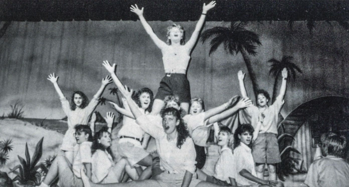 Many females in shorts and white shirts with hands up and palm trees in background