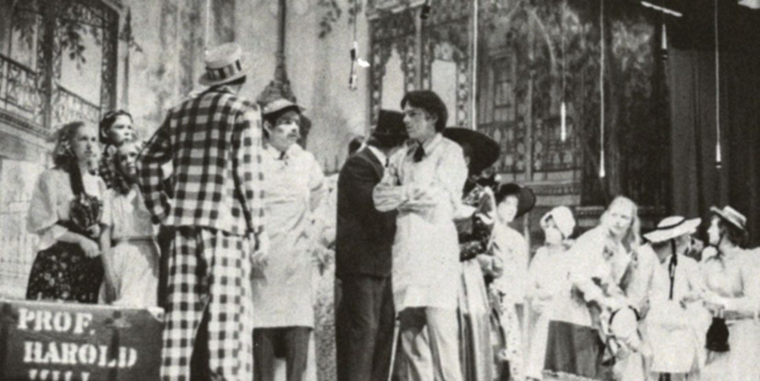 Back of male in plaid suit with hat, and many others on stage as well