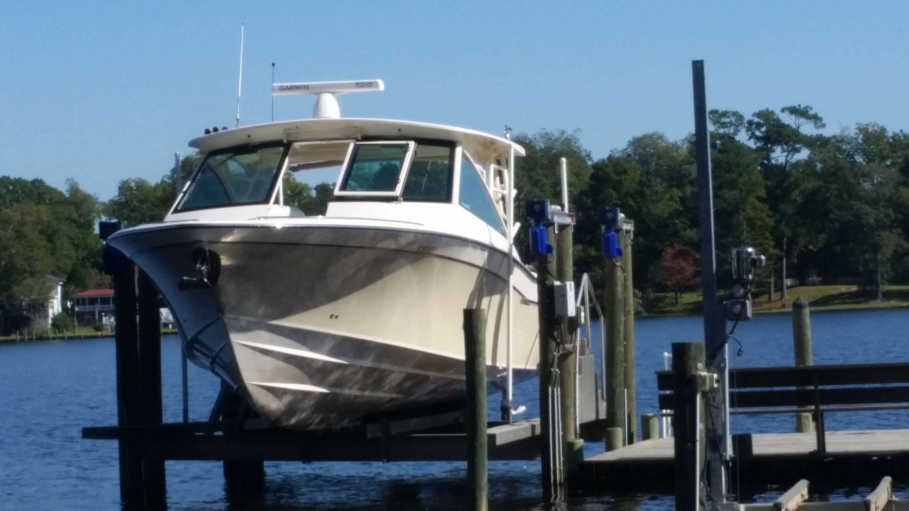 White motor yacht docked at a marina on calm water, with trees and blue sky in the background